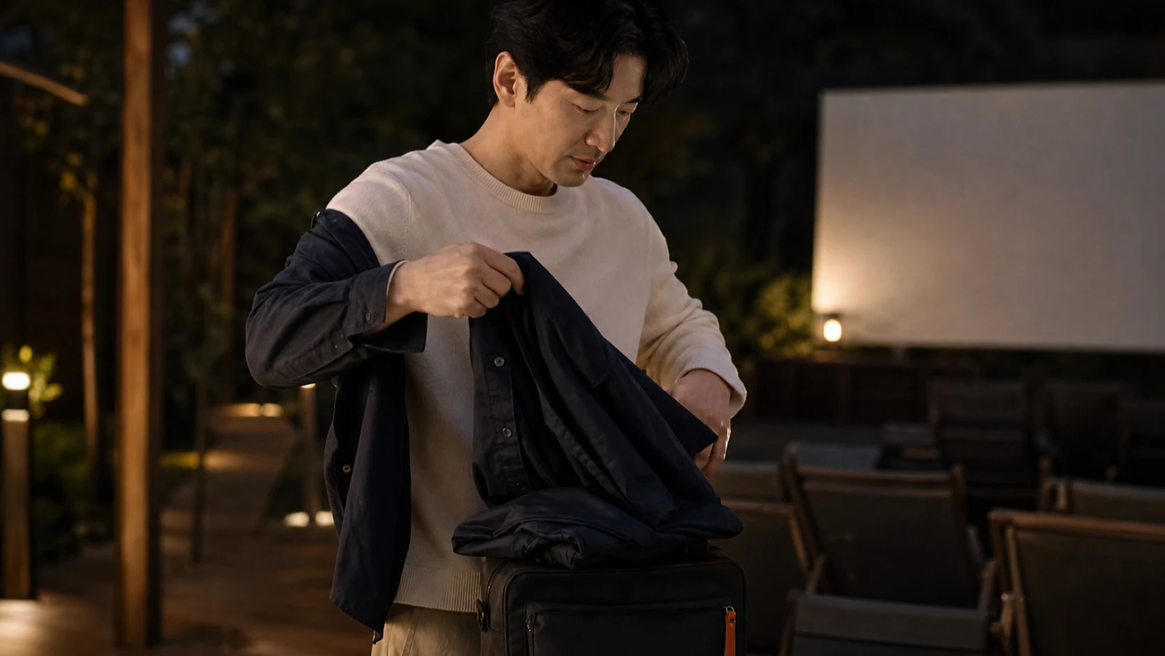 야외 상영 전 가벼운 겉옷을 작은 가방 위에 정리하며 한 벌의 조절 방식을 살피는 남성