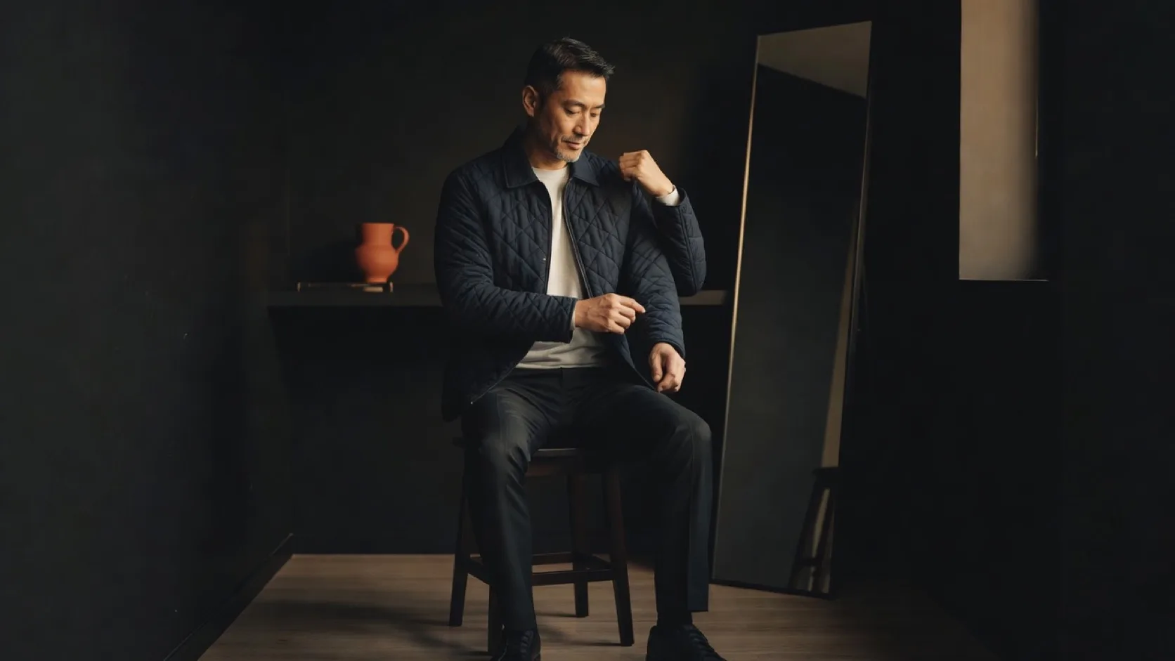 네이비 퀼팅 재킷과 흰 티셔츠, 검은 바지 차림의 남성이 거울 옆 의자에 앉아 한 손으로 반대쪽 어깨를 잡고 소매와 어깨의 여유를 확인하는 장면